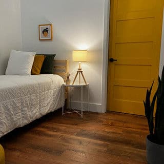 Cozy bedroom featuring a bed, lamp, and yellow door against warm wooden floors.