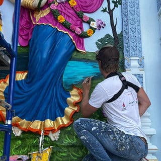 Artist painting a vibrant mural of a goddess near a tranquil water scene.