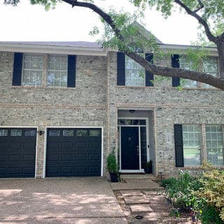 Two-story brick house with dark green shutters and garage doors, surrounded by trees and landscaping.